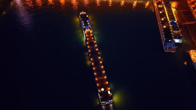 Top down aerial view of cargo barge on Danube river. Overhead drone shot of illuminated vessel on dark water, golden lights, river reflections.