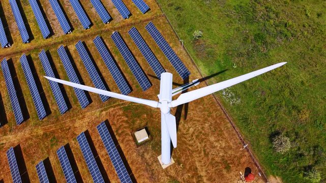 Top down aerial view of wind turbine rotor over solar panels field. White propeller blades casting shadow on blue photovoltaic arrays, orthogonal perspective.
