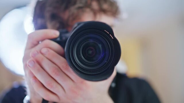 Photographer aiming DSLR camera lens forward, smiling young man home studio.