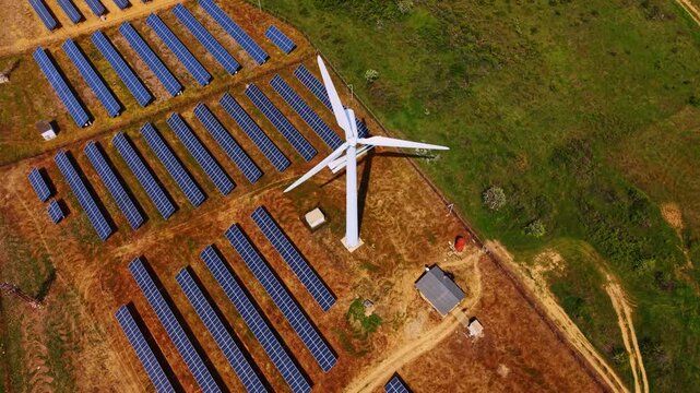 High angle aerial view of wind turbine and solar panel installation. White generator standing among blue photovoltaic modules on brown soil, overhead perspective.