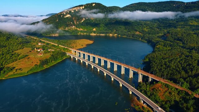 Aerial view of bridges over water near mountain. Top view of parallel railway and road viaducts over blue river near green hills with morning fog