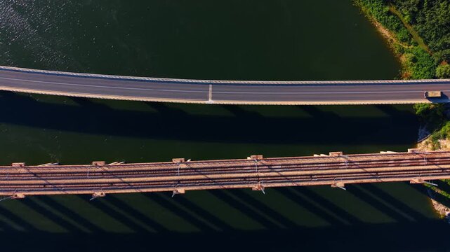 Top down aerial view of parallel road and rail bridges. Direct overhead perspective shows concrete structures casting dark shadows on calm deep water.