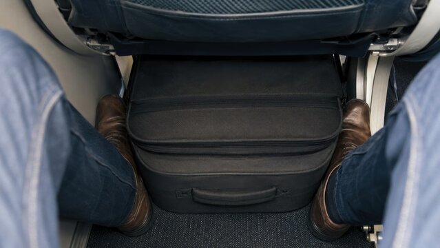Passenger's legs in blue jeans positioned beside a black carry-on bag under an airplane seat, showcasing limited legroom and discomfort during flight