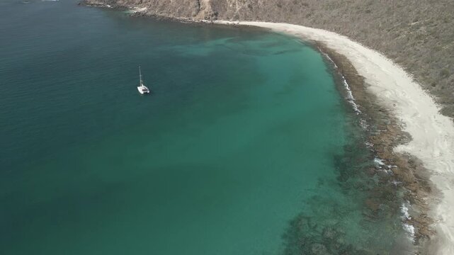 Beautiful aerial drone shot of a remote island cove with a deserted sandy beach beneath high cliffs and a catamaran anchored in turquoise water, filmed in a flat log profile for color grading