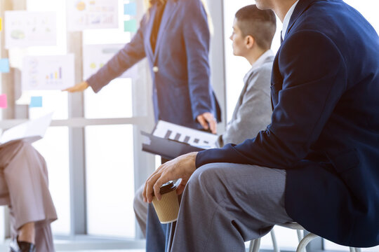 Businessman holding a disposable coffee cup while listening to a female leader presenting business charts on a glass wall during a team strategy meeting in the meeting room at modern office.