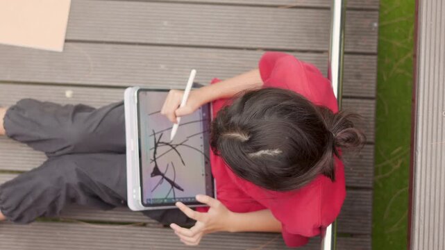 Top view of an Asian kid enjoying a relaxing hobby, drawing digital art on a tablet. Selective focus captures the elementary student spending casual leisure time at an outdoor school.