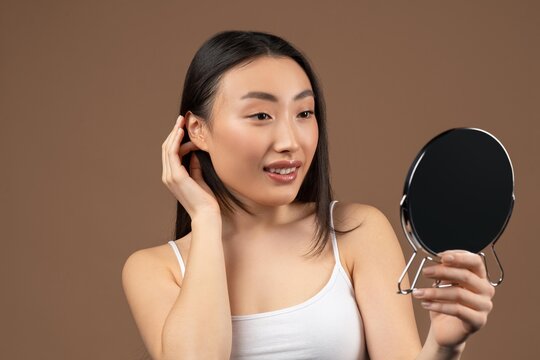 Happy Young Asian Woman Smiling While Applying Moisturizer In Front Of Mirror At Home, Taking Care Of Skin And Enjoying Her Beauty Routine. Relaxing Skincare Concept In Comfortable Environment.