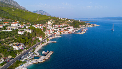 Fototapeta premium High-angle view of a Mediterranean seaside village featuring red-roofed houses, a coastal road, and small boat docks under steep green mountains and clear blue water.