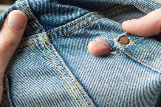 Hands inspect frayed hole near front pocket rivet on blue jeans and show garment wear that calls for repair or sustainable reuse.