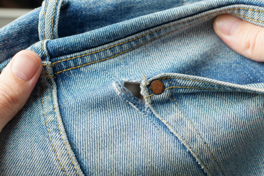 Hands display frayed hole beside front pocket rivet on blue jeans and show worn denim damage that requires careful clothing repair.