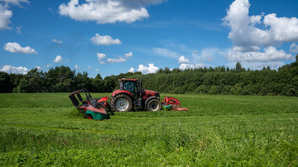 Red tractor mowing a green forage field under a bright blue summer sky © Edijs
