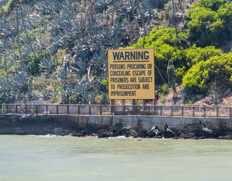 A legal warning sign stands amidst desert cacti on the rugged cliffs of Alcatraz Island overlooking the waters of San Francisco Bay in California.