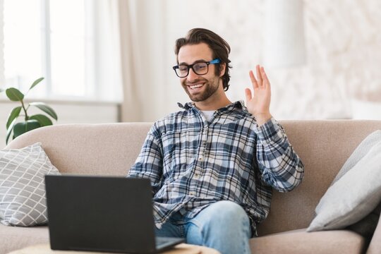 Smiling Caucasian Male In His 30s Video Calling On Laptop While Sitting On Couch In Modern Living Room. Engaging In Online Conversation With Joyful Emotion, Relaxing At Home. Business Concept.
