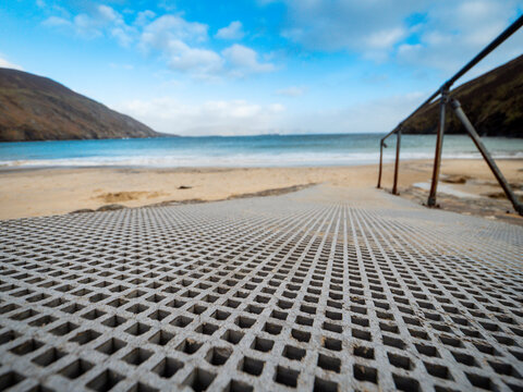 Metal grid walkway leads toward sandy beach. Blue sky with clouds above. Rocky hills surround calm sea. Sand stretches to waterline. Metal railing on right side. No people or animals visible, travel