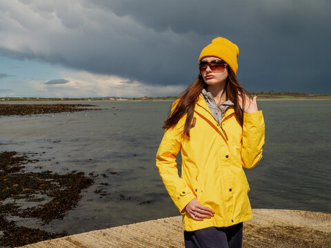 Teenager girl in yellow travel jacket standing on a pier. Huge dark storm cloud in the background. Travel in harsh weather conditions with sun and rain. County Sligo, Ireland.