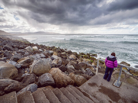 Child in a purple jacket stands on stone steps by a rocky shoreline. Waves crash against large, wet boulders under a cloudy sky. The child looks out at the ocean, Strandhill, Sligo, Ireland. Travel