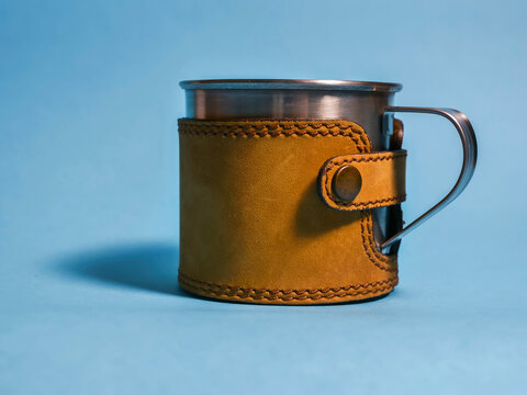 Vintage metal cup wrapped in brown leather sleeve. Leather strap fastened with button. Set against solid blue background. Travel equipment and hand craft.