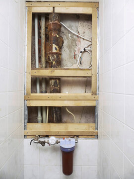 Exposed plumbing and wooden framing in tiled bathroom wall. Rusty pipes and exposed fixtures show renovation progress. Brown water filter and meter attached to wall below frame