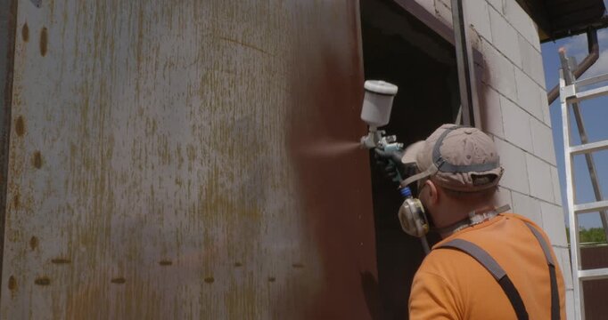 Professional worker in protective gear using a spray gun to apply brown paint on an old, rusty metal surface, demonstrating the process of industrial painting and garage door renovation.