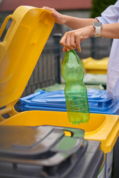 Hand placing empty plastic bottle into yellow recycling bin outdoors