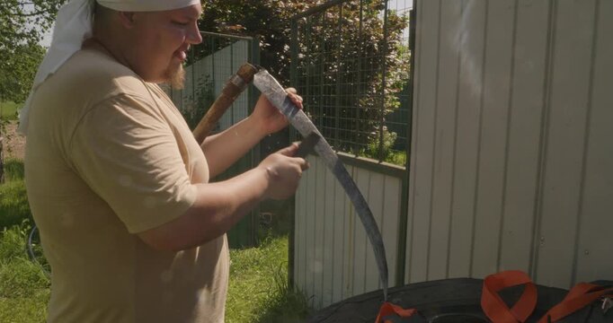 Caucasian farmer preparing for harvest work by sharpening the blade of a traditional scythe with a whetstone and water in his backyard, maintaining the agricultural tool on a sunny day.