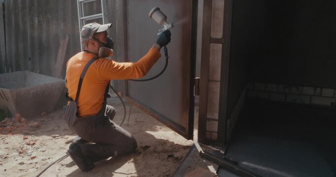 Professional painter in protective overalls and respirator using an airbrush to apply primer on a metal surface, working outdoors at a construction site on a sunny day.