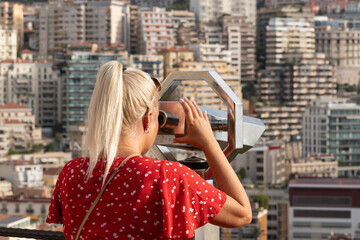 Fototapeta premium Blonde female tourist looking through coin operated binocular telescope at cityscape buildings from viewpoint in Monaco Monte Carlo