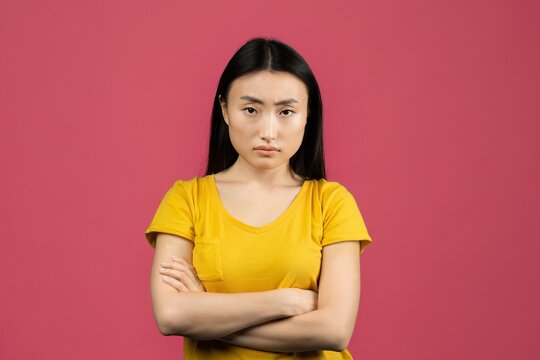Concerned Expression. Serious Young Asian Woman With Black Hair, Crossing Arms And Looking Directly At Camera, Against Vibrant Pink Background. Concept Of Emotions And Feelings.