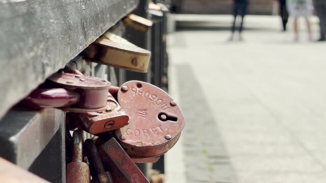 A rusty heart-shaped wedding lock clings to a metal railing. Distant people stand on a blurry walkway in the background. This sweet tradition evokes a deep feeling of romantic nostalgia.