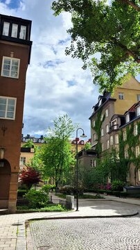 Street view of &Ouml;stermalm district in Stockholm, Sweden, with historic residential buildings, green trees, courtyard gardens and a cloudy blue sky in summer daylight.