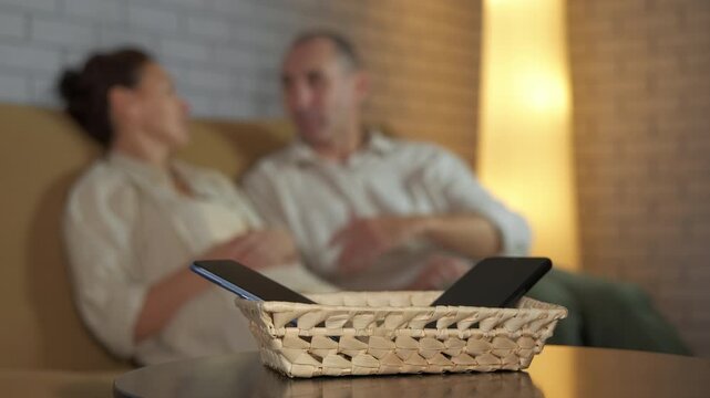 Couple talking together without smartphone distractions. Wicker basket with mobile phones on a table with a blurred couple talking on the sofa in the background, representing a moment of digital detox