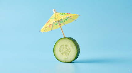 Naklejka premium Fresh green cucumber slice with a small yellow paper cocktail umbrella on a bright light blue background, creative food concept for summer and relaxation