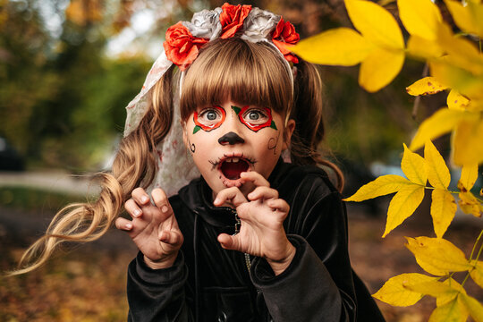 Cute little girl in Halloween face paint with autumn leaves outdoors