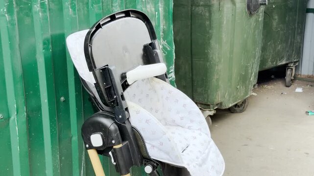 A lonely baby high chair stands abandoned against a green fence. It sits forgotten near large trash dumpsters, creating a sad and melancholic mood of past childhood.
