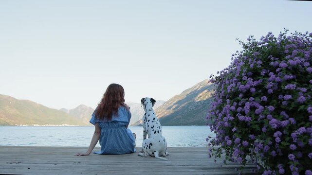 A young girl in a blue dress sits beside a Dalmatian dog on a wooden dock by the water. The two face the lake surrounded by flowers and mountains in soft evening light.