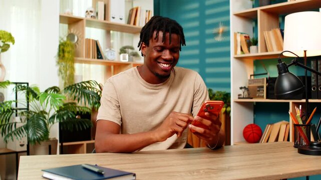 African American young man on home table reads smartphone message with warm smile and feels happy about good news. Black guy on home table checks text chat for social contact fun or nice online news.