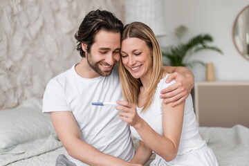 Fototapeta premium Smiling Caucasian Couple, Young Ages, Celebrating Positive Pregnancy Test Together At Home, Sitting On Bed In Modern Bedroom. Emotions Of Joy, Anticipation, And Parenthood Concept.