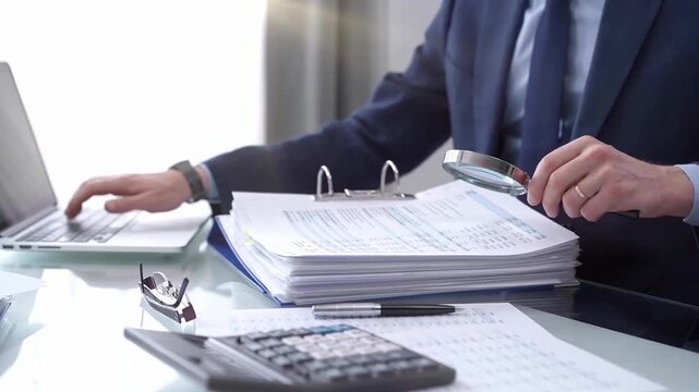 Businessman reviewing ring folder of financial documents with magnifying glass at desk in modern office. Audit and taxes in business