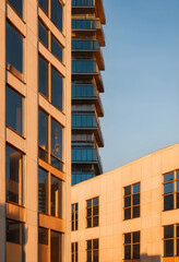 Fototapeta premium Modern urban architecture with glass windows and balconies reflecting Golden hour sunlight against a clear blue sky, Golden hour sunlight illuminates the facades, creating warm reflections