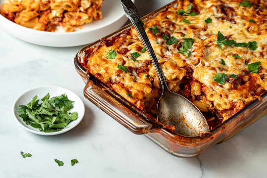 A baking dish of baked ziti hot out of the oven with one serving on a plate.