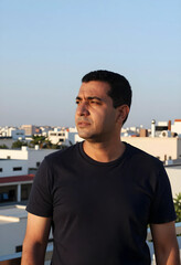 Fototapeta premium Middle eastern man looking away with a serious expression on a rooftop at sunset, urban background, the urban background features city buildings and a clear sky