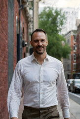 Obraz premium Smiling man in a white shirt and brown pants walking confidently on a city street with brick buildings, the urban background features brick buildings and blurred elements, creating a sense