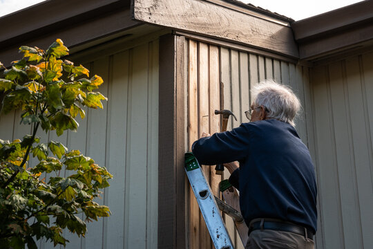 Elderly on an extension ladder using a hammer to nail in new wood siding boards, homeowner maintenance and upkeep, making outdoor repairs on house
