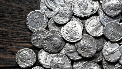 Obraz premium Pile of ancient Roman silver coins on a dark wooden background. Collection of silver denarii featuring portraits of emperors. Historical numismatics and archaeology concept.