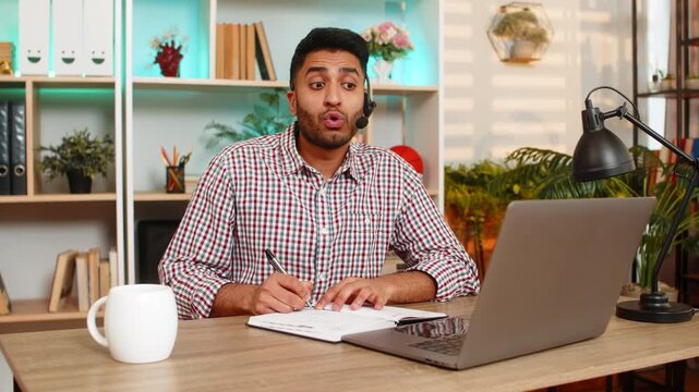 Indian man on home table works in headset as support operator writes notes during remote service call. Arabian guy on home table uses laptop microphone with focused mood for online help job indoors.