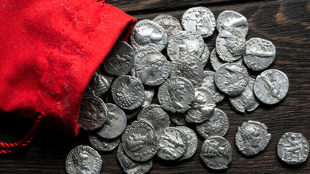 Ancient silver coins spilling from a red cloth pouch onto a dark wooden table. Roman denarii treasure hoard. Numismatics and historical wealth concept