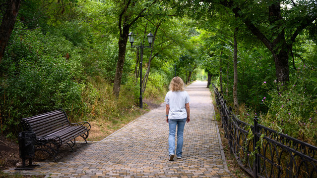 Woman walking away on a paved path in a green park. Back view of a blonde female person in casual clothes strolling outdoors. Summer leisure activity in nature