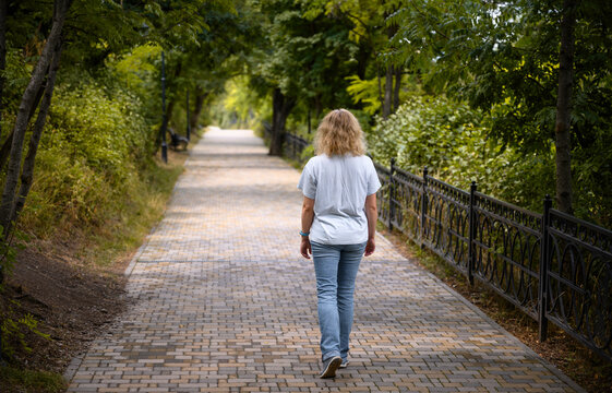 Back view of a lonely young woman walking on a paved path in a green park. Adult female person strolling alone through a tree lined walkway in summer