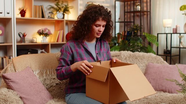 Young woman on home sofa opens delivery parcel then frowns at wrong order after online purchase surprise. Adult girl on home sofa shows shock and stress over return refund or shopping mistake at home.