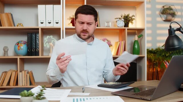 Young man on home table checks bills with tense lips and helpless hands after unexpected payment shock. Adult guy on home table tries to review costs and manage budget stress during home money crisis.